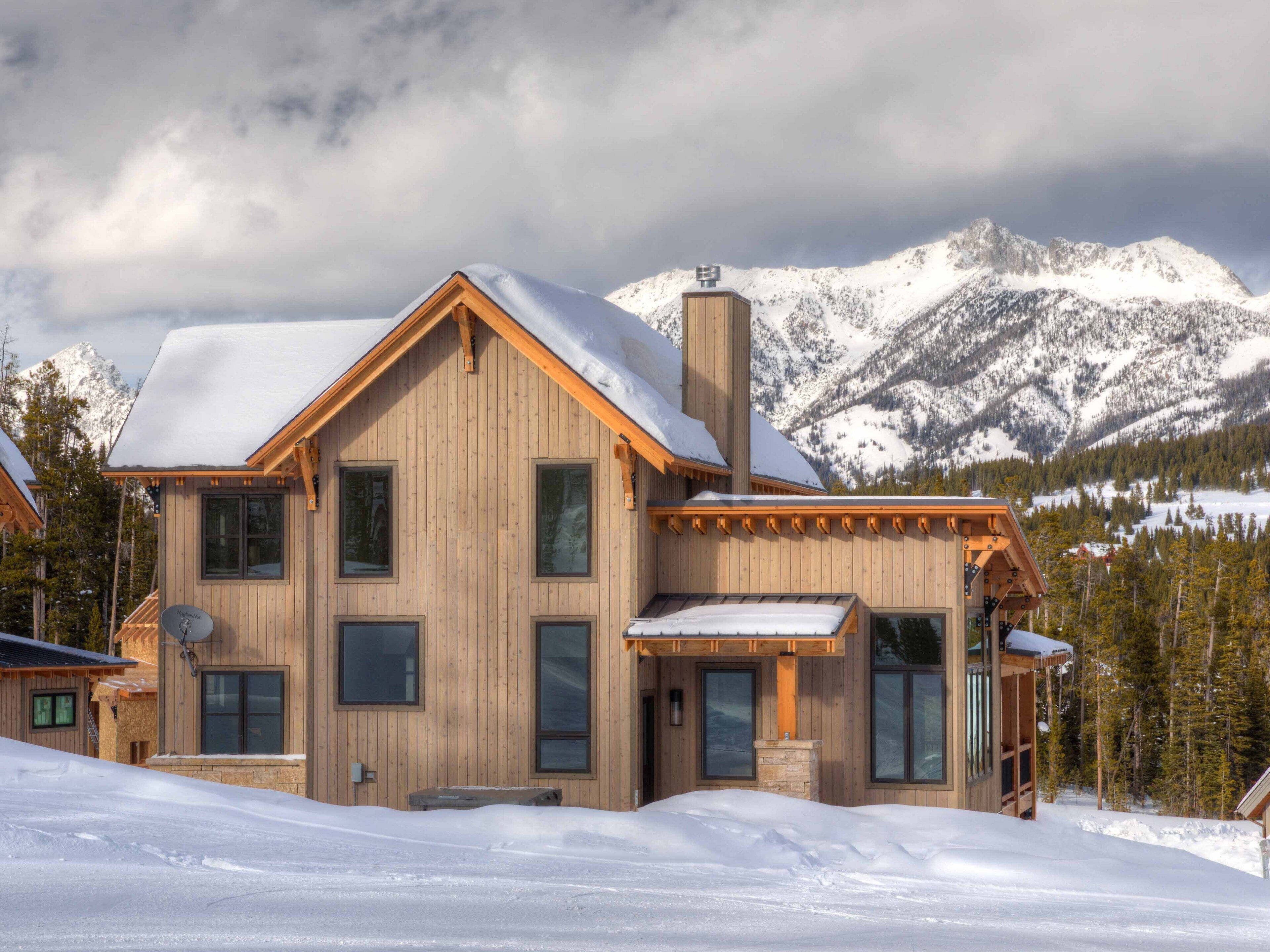 A wooden cabin with a large chimney, multiple windows, and a snow-covered roof, set against a mountainous backdrop.