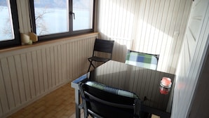 Una habitación pequeña con piso de madera, una ventana con vista a un lago, una silla negra, una mesa de vidrio y una vela roja.