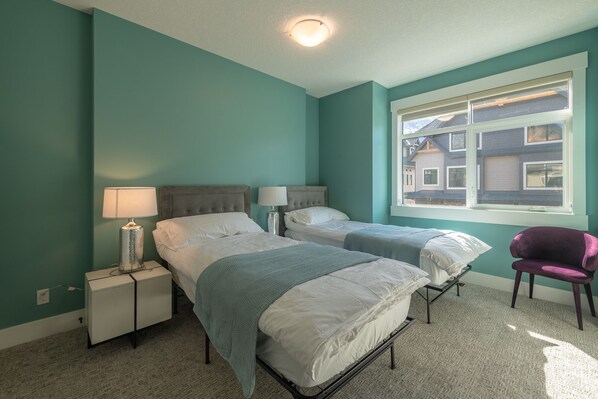A bedroom with two beds, a nightstand, a lamp, a chair, and a window with a view of buildings.