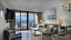 Un vaste salon doté d’une grande fenêtre offrant une vue sur la mer, d’un téléviseur à écran plat fixé au mur, d’un canapé avec deux lampes et d’un coin repas avec des chaises.