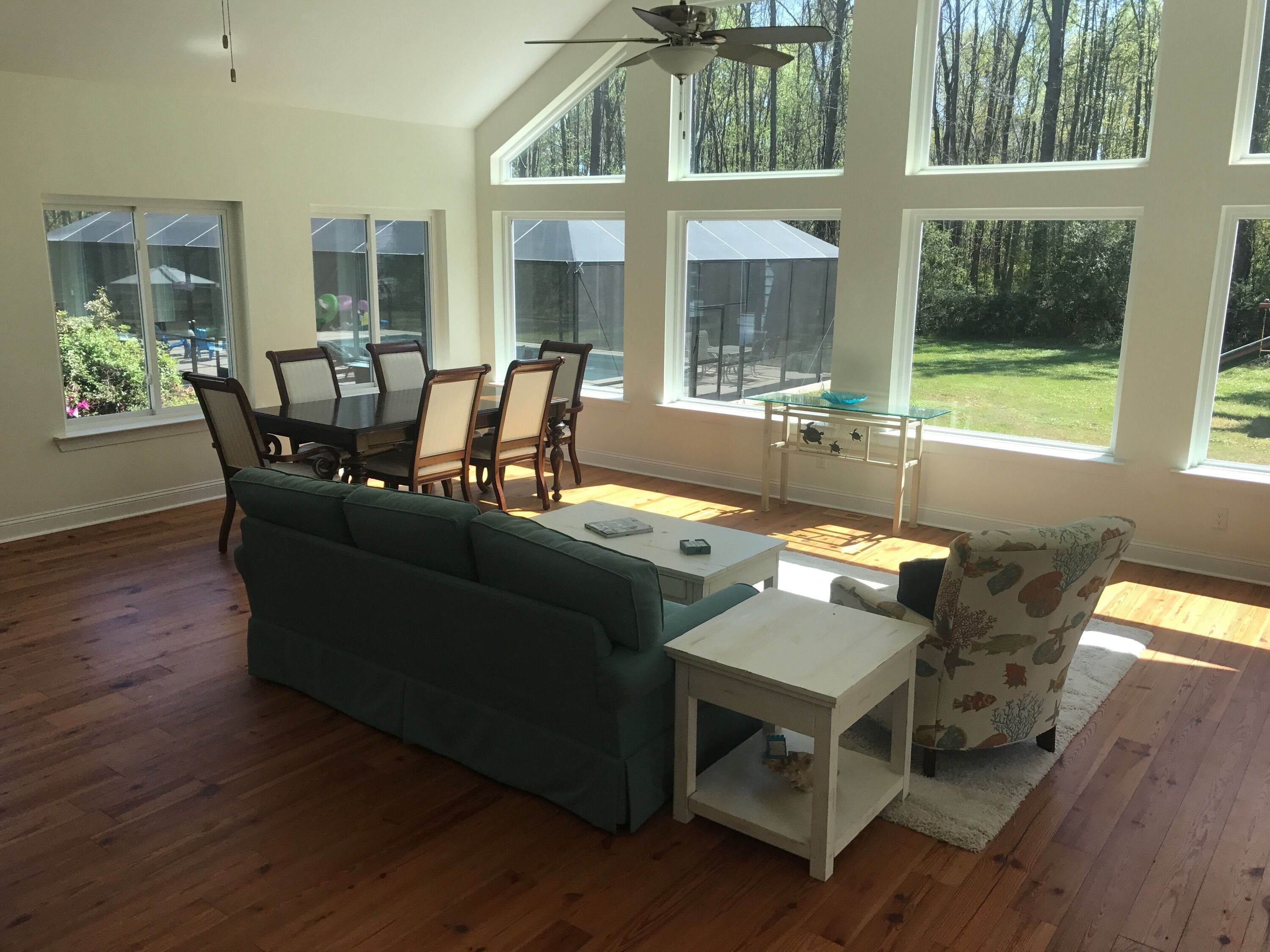 A spacious living room with a wooden floor, a green sofa, a white coffee table, and a patterned armchair. Large windows offer views of a garden and a building with a glass roof.