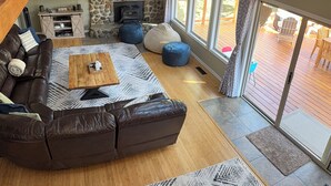 A modern living room with a stone fireplace, a large brown leather sofa, a wooden coffee table, and a large window overlooking a deck.