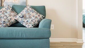 A teal sofa with patterned cushions in a living room with wooden flooring and a plain wall. - Cute Condo in Rock Hill-45 (Rock Hill)