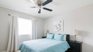A bedroom with a bed, nightstands, a ceiling fan, a window with curtains, and a wall-mounted lamp. - Cute Condo in Rock Hill-45 (Rock Hill)