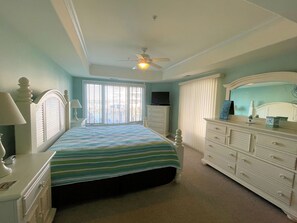 Une chambre à coucher comprenant un lit, une commode, une table de chevet, une lampe, un ventilateur de plafond et une fenêtre avec des stores.