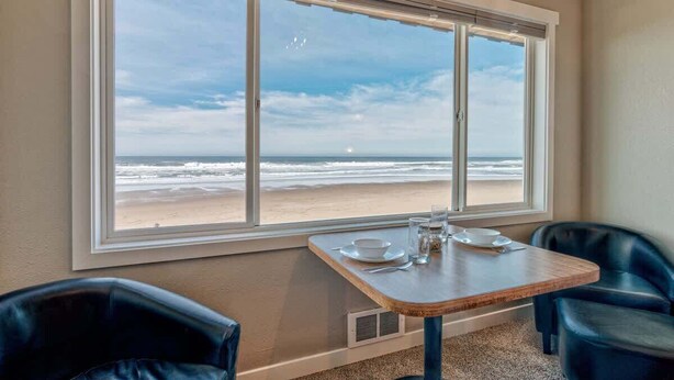 Ein Zimmer mit großem Fenster und Blick auf einen Strand, ein Tisch gedeckt mit Tellern und Gläsern, sowie zwei schwarze Ledersessel.