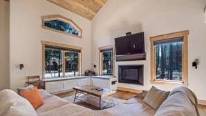 Ein gemütliches Wohnzimmer mit einer großen Couch, einem Kamin und bodengrossen Fenstern mit Blick auf den Wald.