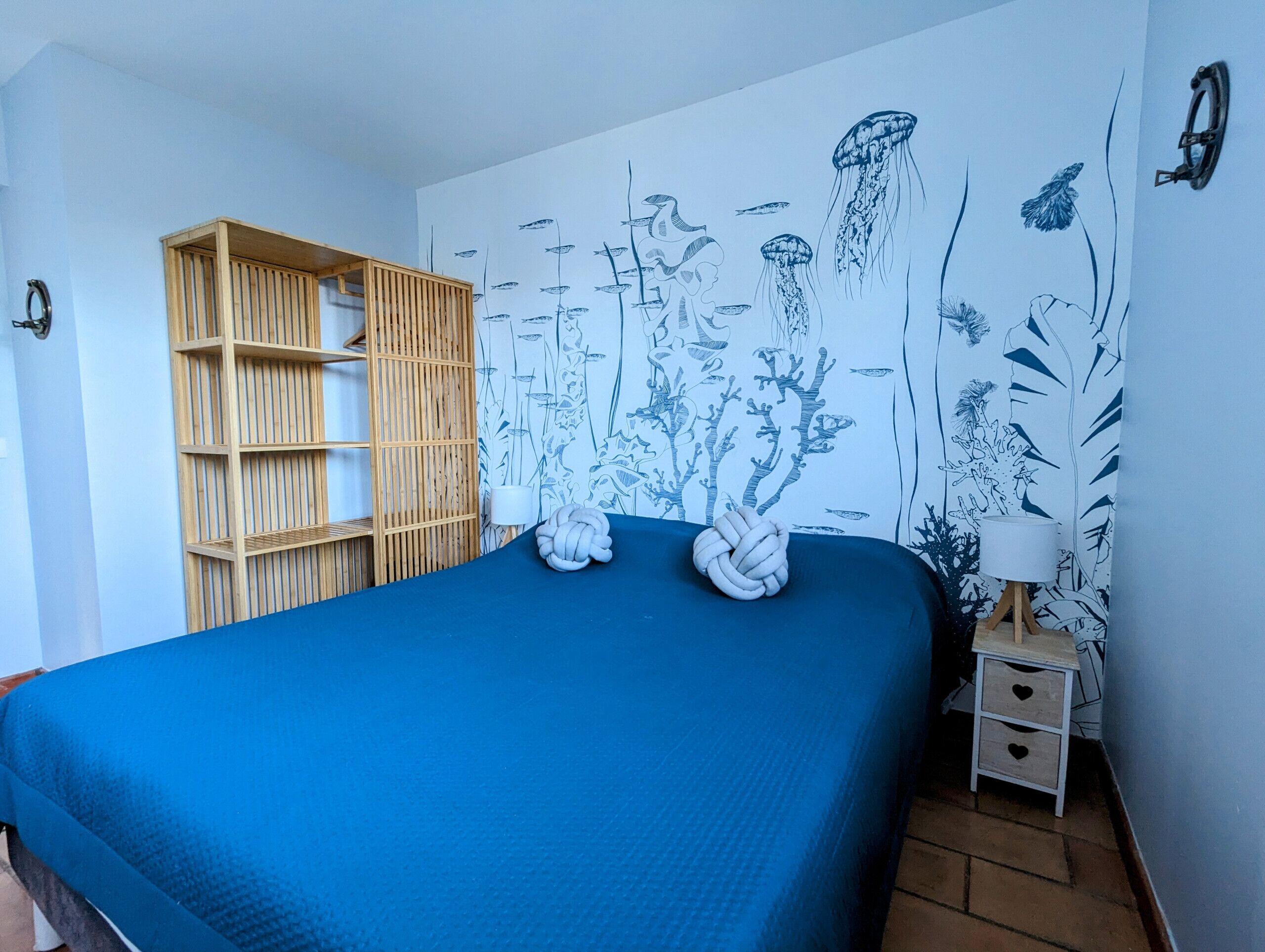 A bedroom with a blue bedspread, a wooden shelf unit, and a wall mural of marine life.