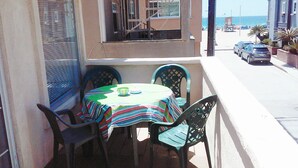 Une petite table d’extérieur, recouverte d’une nappe colorée, entourée de quatre chaises, donnant sur une rue bordée de voitures garées et offrant une vue sur l’océan.