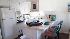 Une cuisine avec des meubles blancs, une cuisinière, un four à micro-ondes et un évier. On y trouve une poubelle et un petit îlot central avec des chaises et un plateau.
