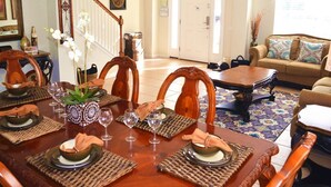 A dining area with a wooden table set for four, surrounded by chairs. There is a staircase leading to another level, and a sofa in the background.