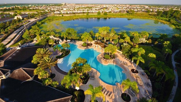 A resort with a large central pool surrounded by palm trees and a view of a residential area in the background.