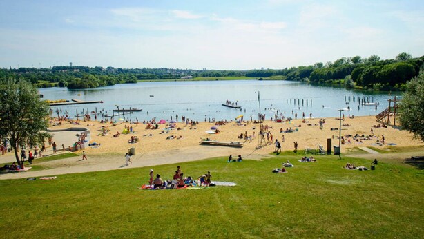 Een strand met een meer, mensen ontspannen en boten.