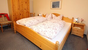 Un dormitorio con cama y armario de madera, una silla, una mesita de noche con lámpara y un cuadro en la pared.