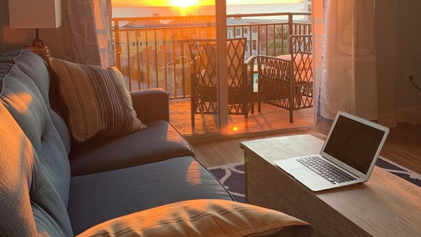 A living room with a sofa, a laptop on a table, and a view of the sunset through a balcony. - Gulf View Condo, Sleeps 6, Pool, Steps to Beach (Madeira Beach)