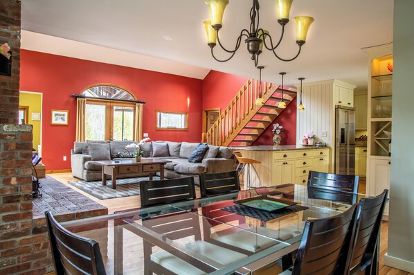 Un salon avec un mur rouge, un escalier en bois, un coin repas avec une table en verre et une cuisine avec des meubles blancs.