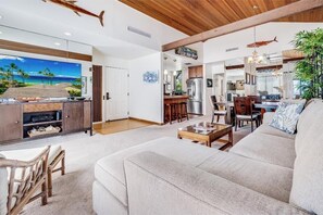 A spacious living area with a large sofa, wooden coffee table, and a dining area with chairs. A kitchen with modern appliances is visible in the background.