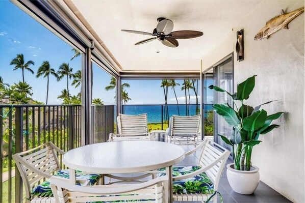 A balcony with a round table and chairs, a ceiling fan, and a view of the ocean with palm trees. - Unforgettable Maui Escape: Experience Wailea Elua 1402, Your Oceanfront Haven! (Wailea, Maui)