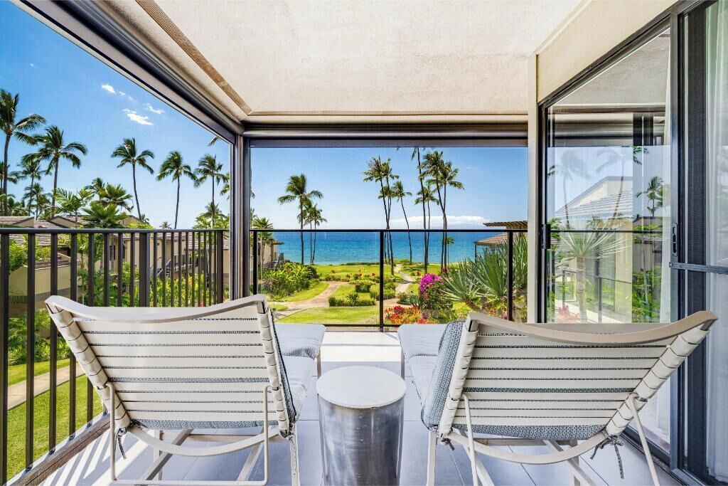 A balcony with two chairs and a table, overlooking a tropical landscape with palm trees and a view of the ocean.