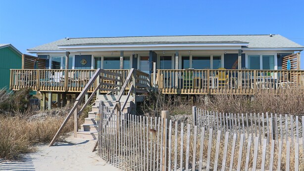 A beachfront house with a wooden deck, a staircase leading to a balcony, and a clear blue sky. - Bright 3BR Beach Home with Deck & Ocean Views (Emerald Isle)