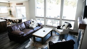 Un salon moderne avec un canapé en cuir marron, un fauteuil blanc et une table basse en bois. Une grande fenêtre offre une vue sur l’extérieur.