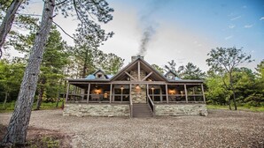 A cabin with a stone exterior, a chimney emitting smoke, and a porch with lights. - Choctaw Wind: Luxurious, Outdoor Furniture, Shuffleboard, Large Hot Tub🛋️🛁🔥 (Broken Bow)
