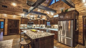 A kitchen with wooden walls, a granite island, stainless steel appliances, and bar stools. - Choctaw Wind: Luxurious, Outdoor Furniture, Shuffleboard, Large Hot Tub🛋️🛁🔥 (Broken Bow)