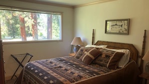 Een slaapkamer met een bed, een raam met uitzicht op bomen, een lamp en een lijst met een afbeelding aan de muur.
