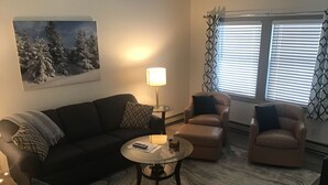 Ein Wohnzimmer mit einem dunkelgrauen Sofa, zwei Sesseln, einem Couchtisch und einer Stehlampe. An der Wand hängt ein Gemälde und ein Fenster ist mit Jalousien verstellt.