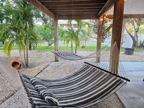 Dos hamacas bajo una pérgola de madera con suelo de grava, rodeadas de palmeras y una valla blanca.