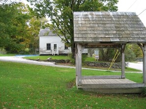 A gazebo with a sloped roof and a wooden deck, surrounded by a grassy area and a stone wall. - Classic Green Mountain Cabin (Mt Holly)
