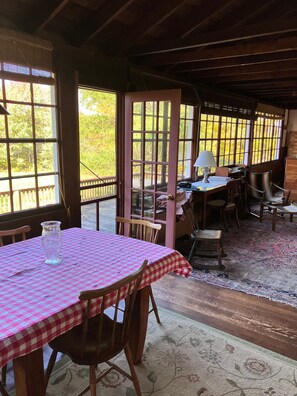 A dining area with a table set for two, wooden chairs, a patterned rug, and a view of trees through large windows. - Classic Green Mountain Cabin (Mt Holly)