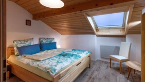 Une chambre avec un plafond en bois, une lucarne, un lit recouvert d’une couvre-lit à motifs floraux, une table de chevet et une chaise.