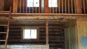 Une cabane en bois avec une chambre à coucher située en mezzanine, une chaise et une fenêtre.
