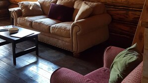 L’intérieur d’un chalet accueillant, avec un plafond en bois, un canapé beige, un fauteuil à motifs rouges et une petite table sur laquelle se trouvent un bol et un livre.