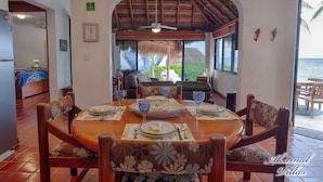 Un comedor con una mesa redonda dispuesta para cuatro personas, rodeada de sillas con tapicería floral. Se observa una estructura de techo de paja al fondo, y la habitación tiene vista al océano.