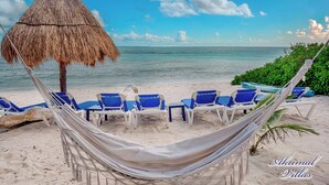 Una playa con una hamaca, sillas reclinables azules y un toldo de paja.