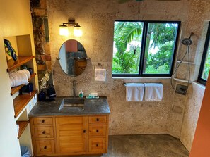 A bathroom with a stone wall, wooden vanity, a round mirror, a window with a view of greenery, and a shower area. - Into the Mystic Lower Suite with POOL and VIEWS! (Coral Bay)