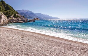 Uma praia de seixos com águas azuis cristalinas e montanhas ao fundo.