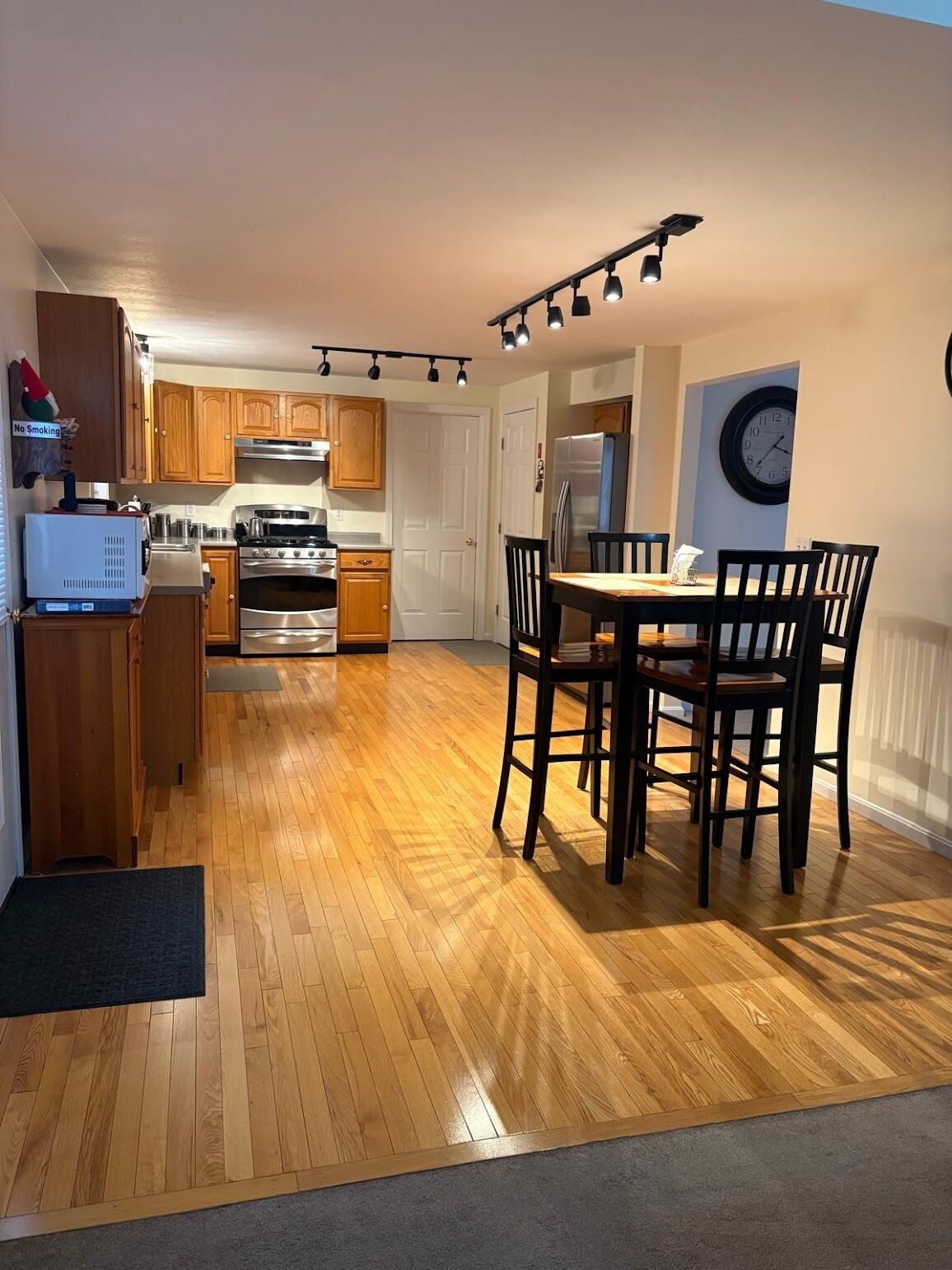 A modern kitchen with wooden cabinets, a dining table with chairs, a refrigerator, and a clock on the wall.