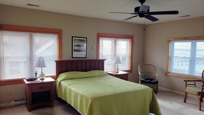 A bedroom with a bed, two nightstands, a chair, a ceiling fan, and large windows with blinds.