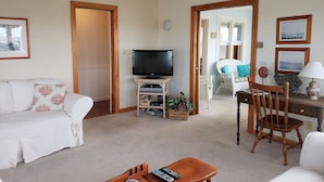 A living room with a television, a white sofa, a wooden chair, and a small table with remote controls.