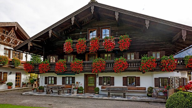 Un edificio tradicional de madera con balcones decorados con flores rojas, un sendero de piedra y un área verde.