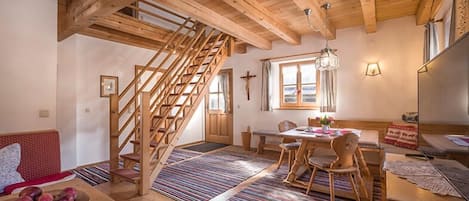 Een gezellige kamer met een houten trap, een zithoek met een tafel en stoelen, en een raam met gordijnen.