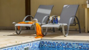 Ein Poolbereich mit Liegestühlen und einem Tisch, ein mit Fliesen ausgelegter Pool und ein Gebäude im Hintergrund.