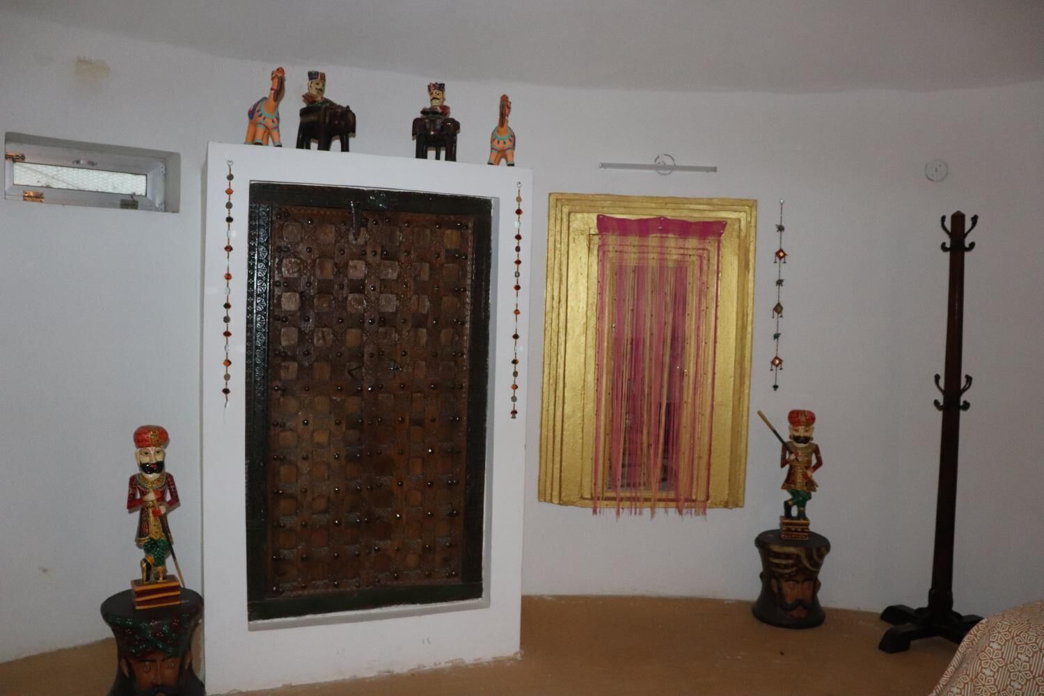 A room with a large wooden door, a window with a pink curtain, and a coat rack.