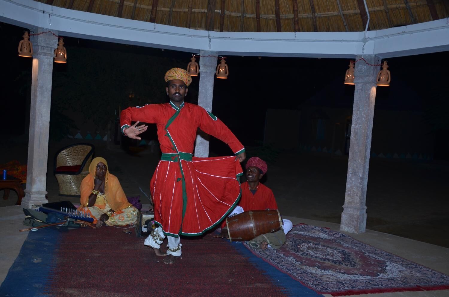A traditional performance with a performer in red attire under a thatched roof structure.
