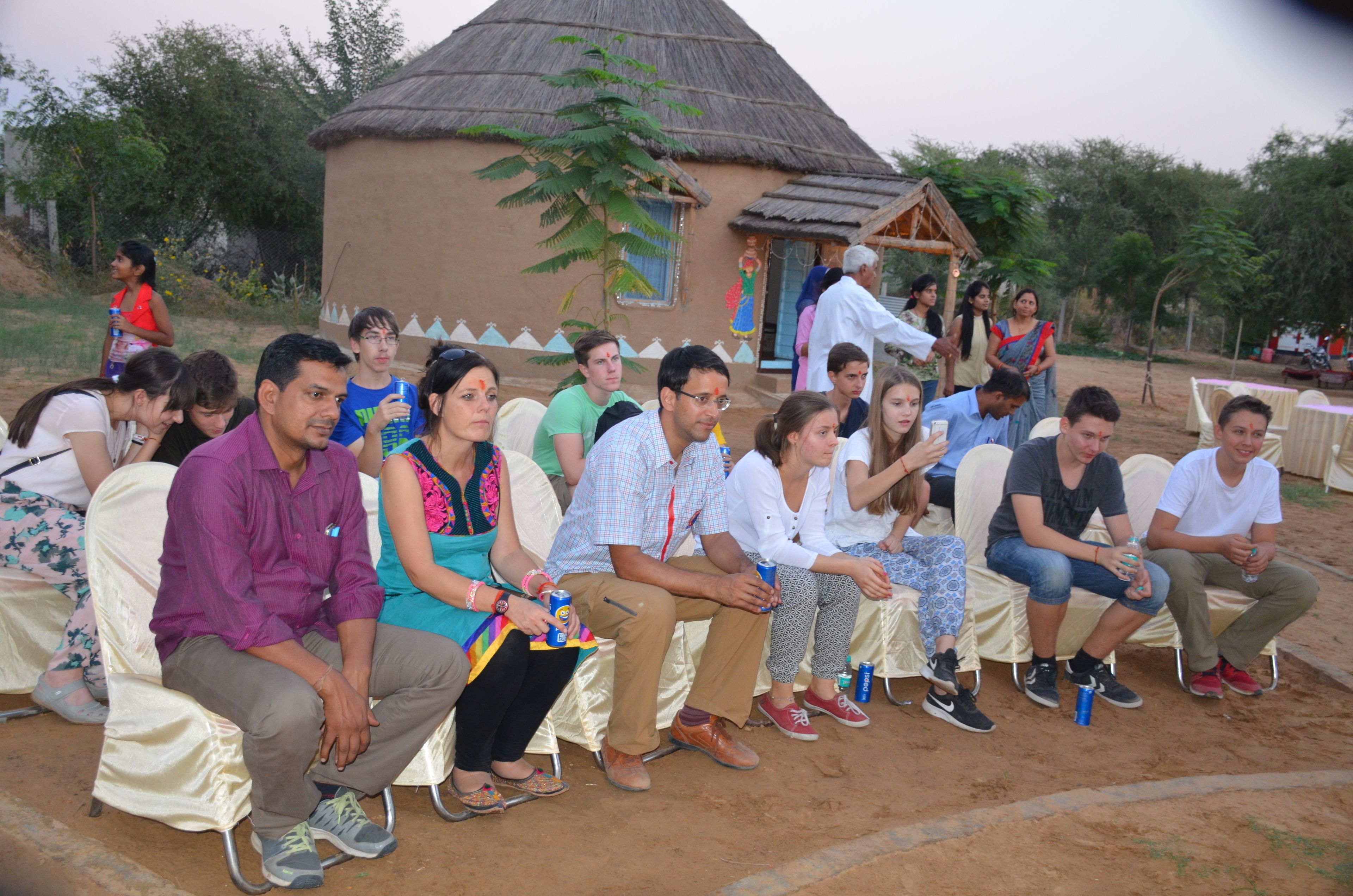 Un grupo de personas sentadas al aire libre en un ambiente semi-formal, con una estructura de techo de paja al fondo.