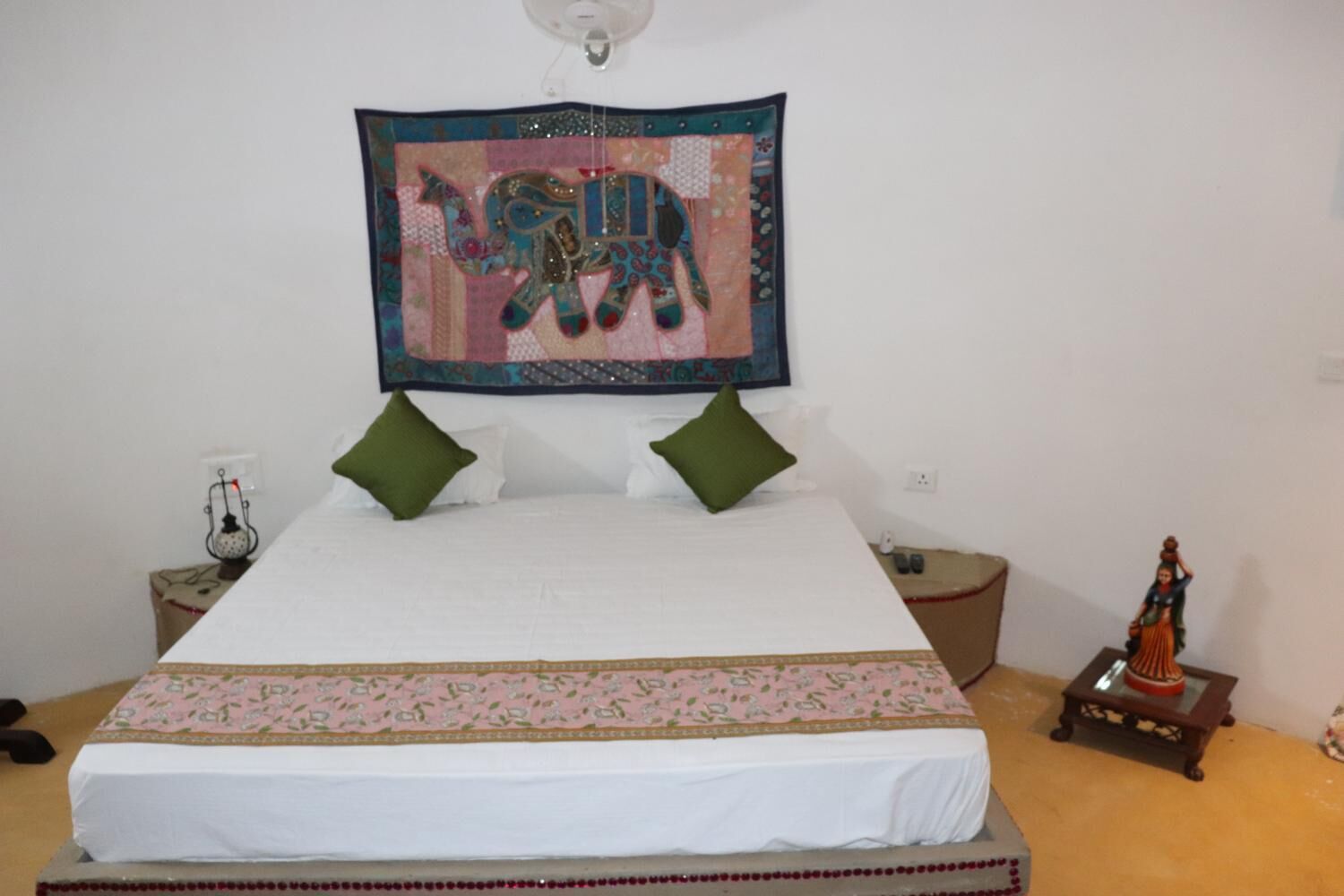 A bed with a white bedspread, green pillows, and a decorative border. A tapestry with an elephant design hangs above the bed.