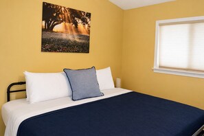 Una cama con una colcha azul oscuro, almohadas blancas y una sola almohada azul. En la pared sobre la cama hay un cuadro con un árbol y un amanecer.